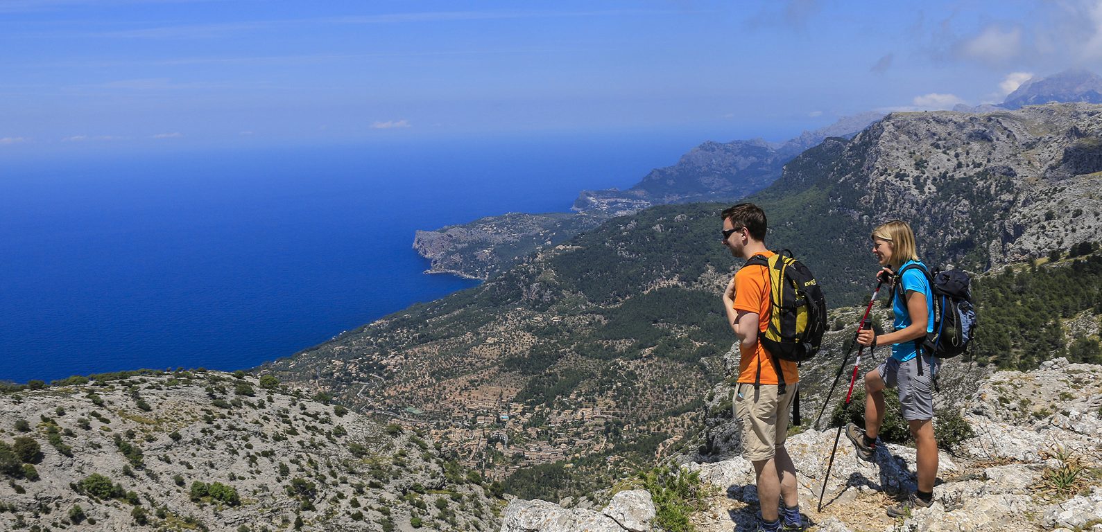 Ett par vandrare blickar ut över kargt medelhavslandskap med blå himmer och blått hav i horisonten.