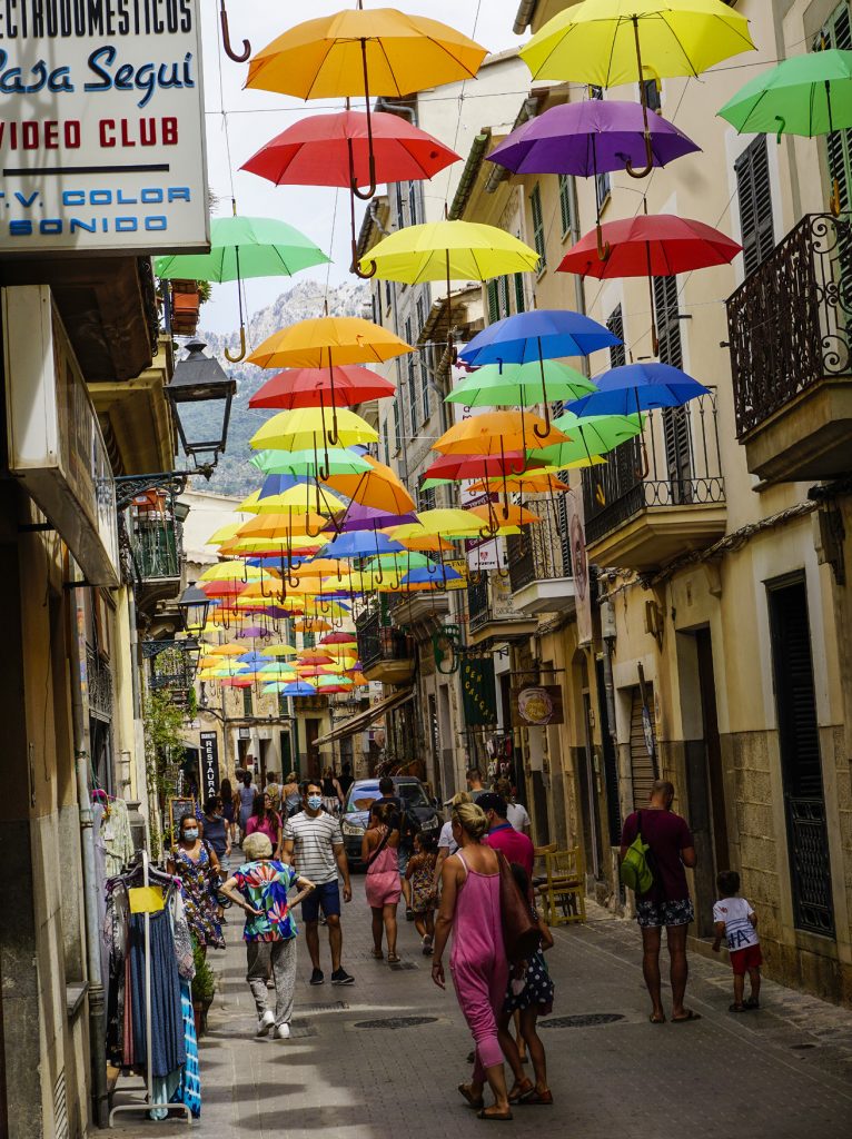 Smal gränd med mycket människor och med färgglada uppspända paraplyer högt upp mellan husen som dekoration.