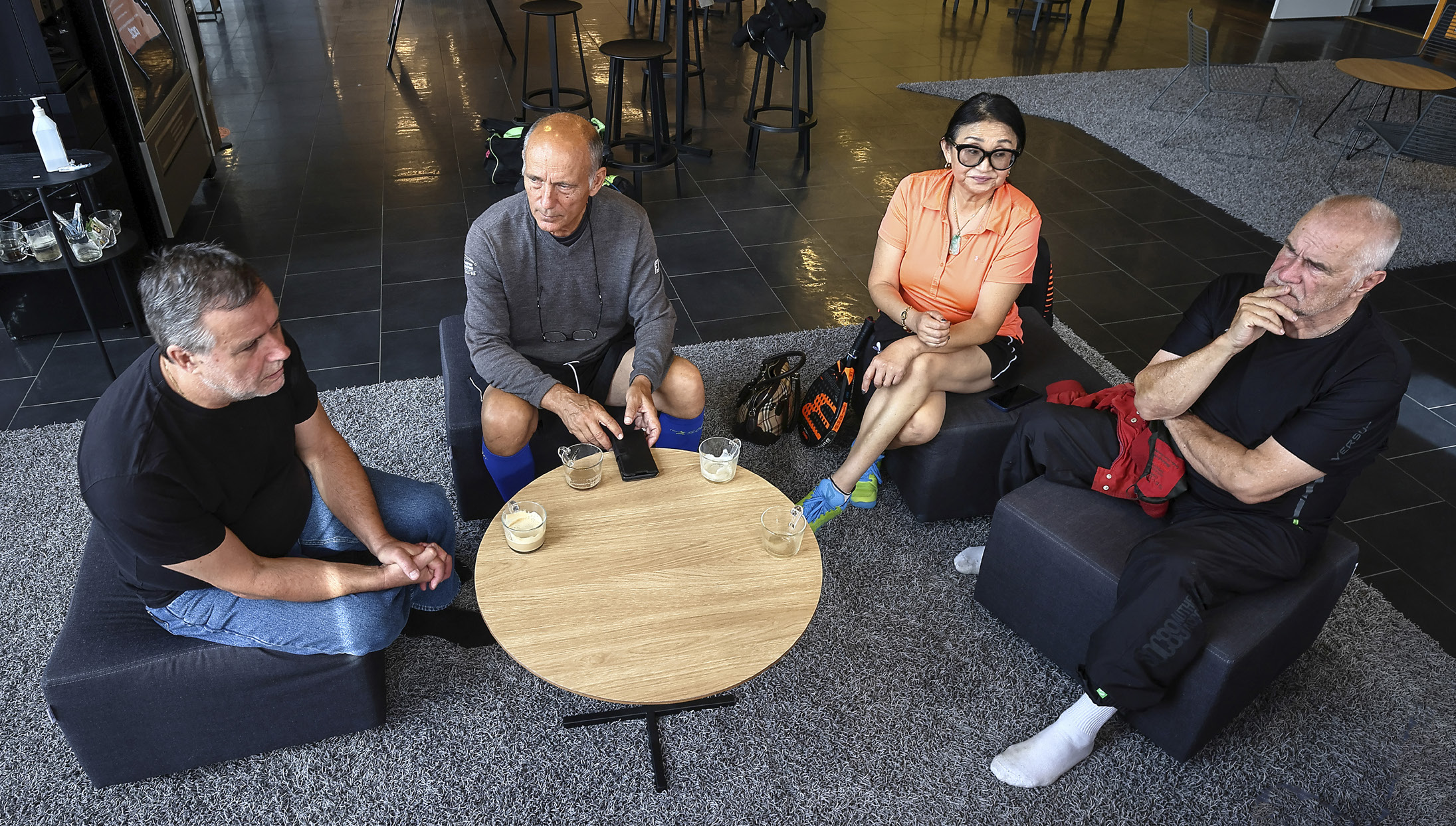 Fyra personer fotograferade ovanifrån, sitter klädda i sportkläder i fåtöljer runt ett runt bord med kaffekoppar.