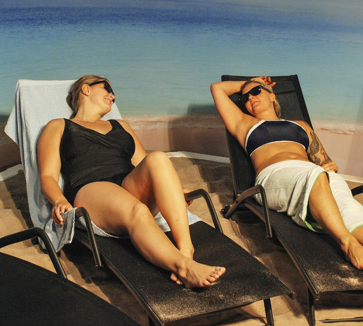 Två kvinnor i baddräkt ligger på solstolar invid en pool.