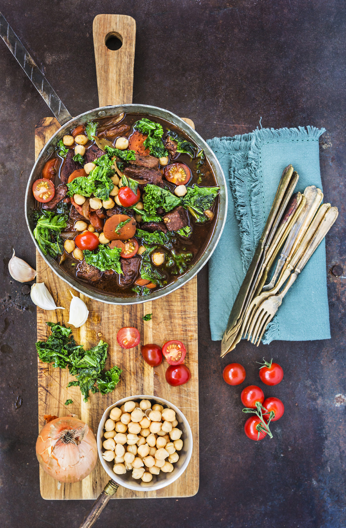 Gryta full med kött och grönsaker står på en skärbräda tillsammans med en liten kastrull kikärter, tomater, bestick, servetter m m. 