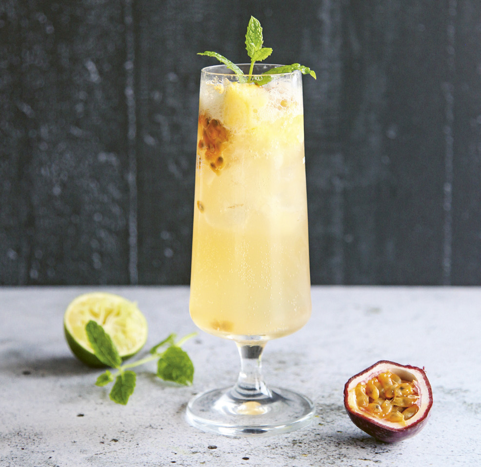 Gulbeige drink i högt cocktailgals, med en halv lime och en halv passionsfrukt på bordet.