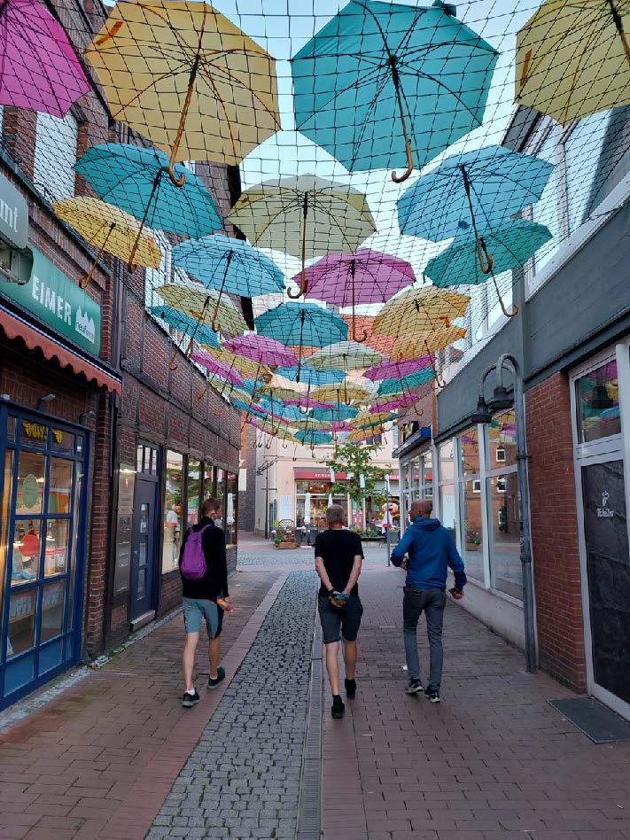 tre killar i "vanliga kläder" fotograferade bakifrån när de går längs en gågata med butiker. På ett nät ovanför deras huvuden ligger en massa öppna paraplyer som dekoration.