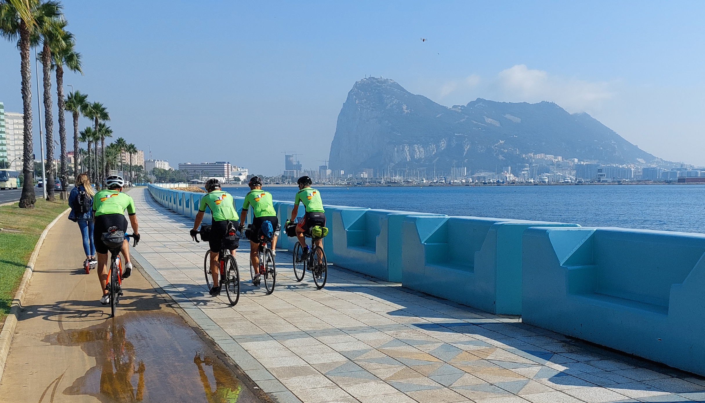fyra cyklande killar i gröna tröjor plåtade bakifrån på en strandpromenad. Gibraltar-klippan tornar upp sig i bakgrunden.