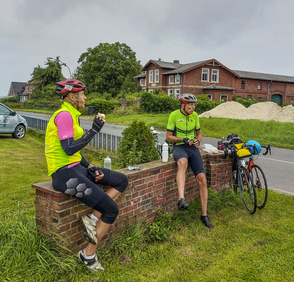 två killar i gröna cykeltröjor sitter på en låg mur och fikar. cyklarna står parkerade lutade mot muren.