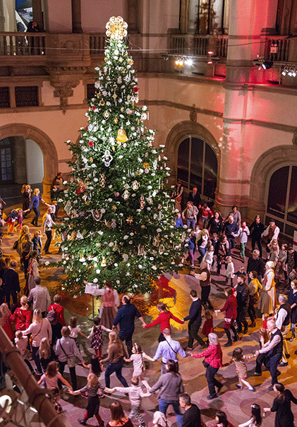 Bild på julfirande människor som dansar runt en dekorerad julgran.